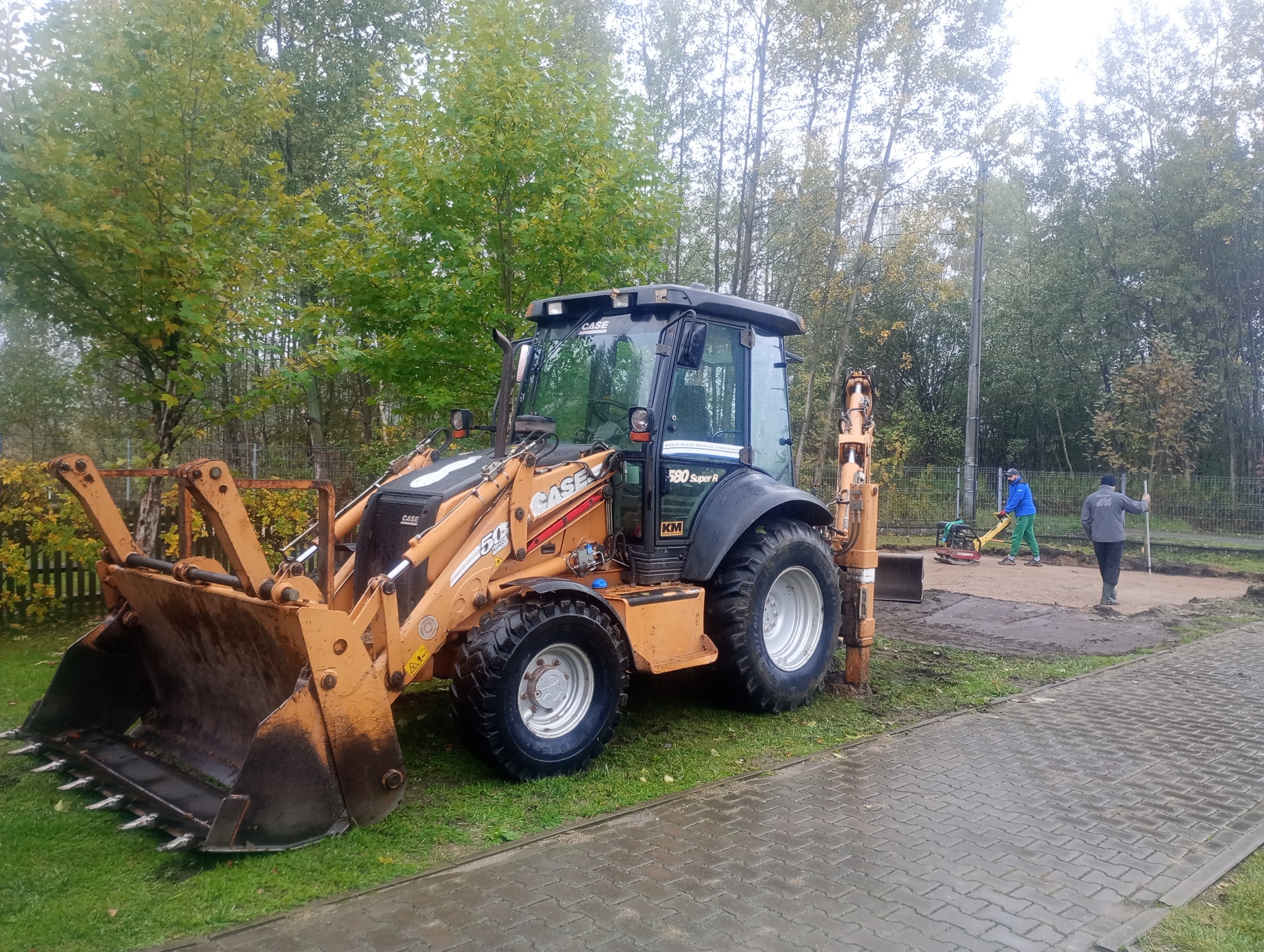 Żółta koparko-ładowarka CASE 580 Super R na trawie, z pracownikami i ubijarką w tle, sugeruje przygotowanie terenu pod budowę. Widok z boku, pochmurny dzień.