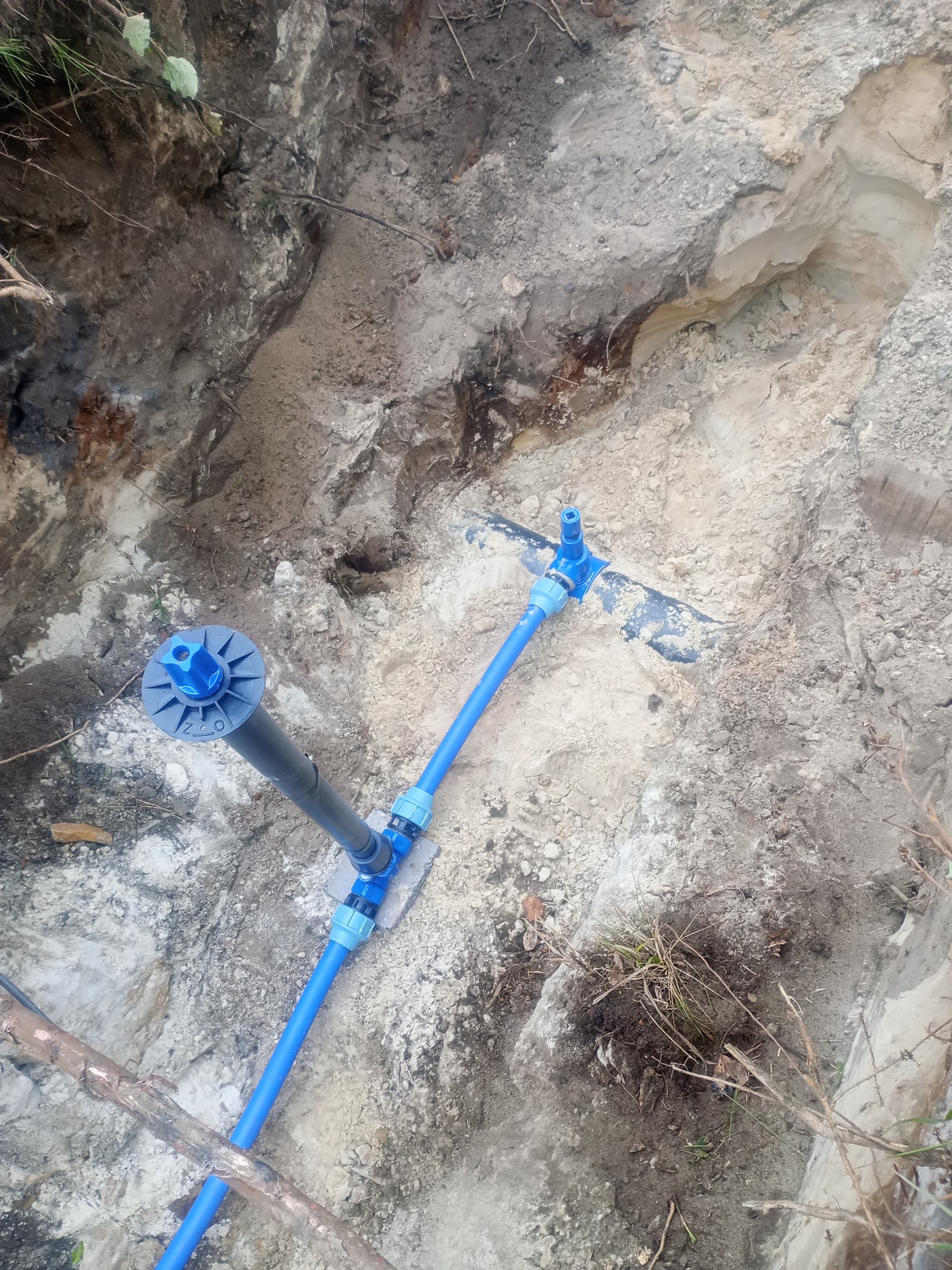 Niebieskie rury instalacji sanitarnej w wykopie ziemnym, widoczne złącza i pionowa rura szara z niebieskim elementem. Wykop w ziemi piaszczystej i gliniastej.