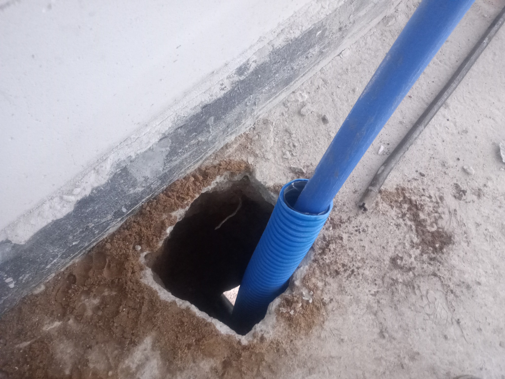 Niebieska karbowana rura w kwadratowym otworze w posadzce, blisko ściany. Widoczny fragment metalowego pręta na betonowej powierzchni.