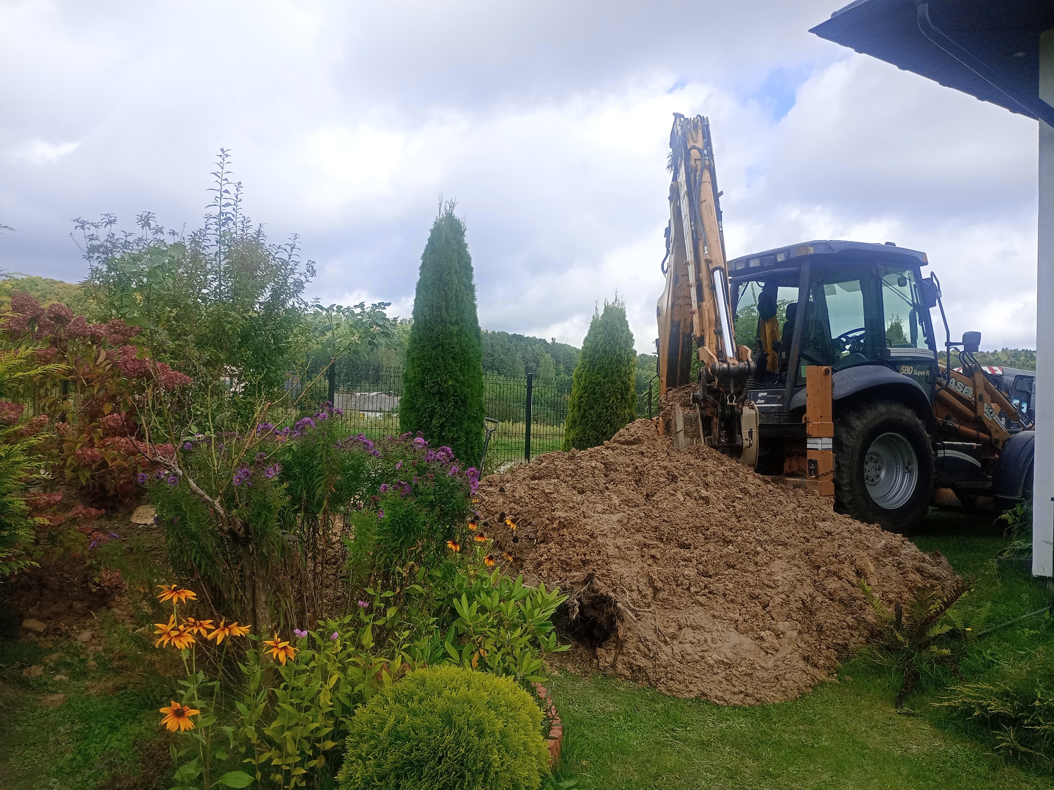 Koparko-ładowarka CASE 580 Super R obok wykopanej ziemi w ogrodzie z kwiatami i tujami, pod pochmurnym niebem. Widoczny fragment dachu budynku.