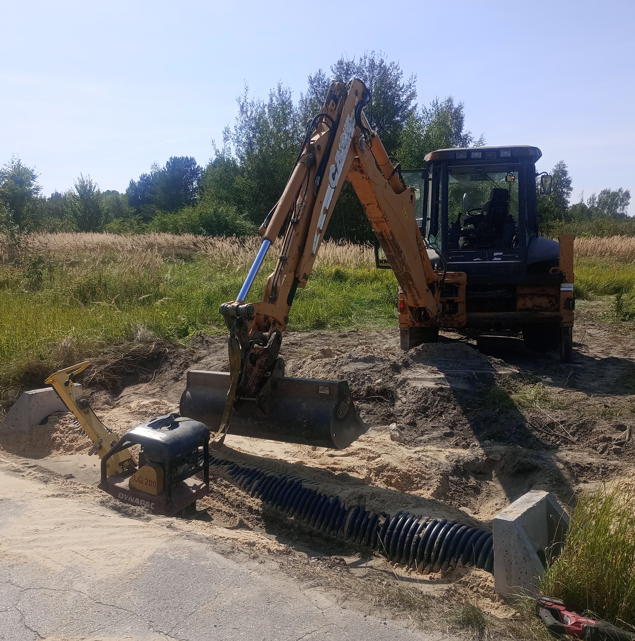 Pomarańczowa koparko-ładowarka Case podczas układania rury drenażowej w piasku, obok ubijarka Dynapac LG 200. W tle zieleń i błękitne niebo.