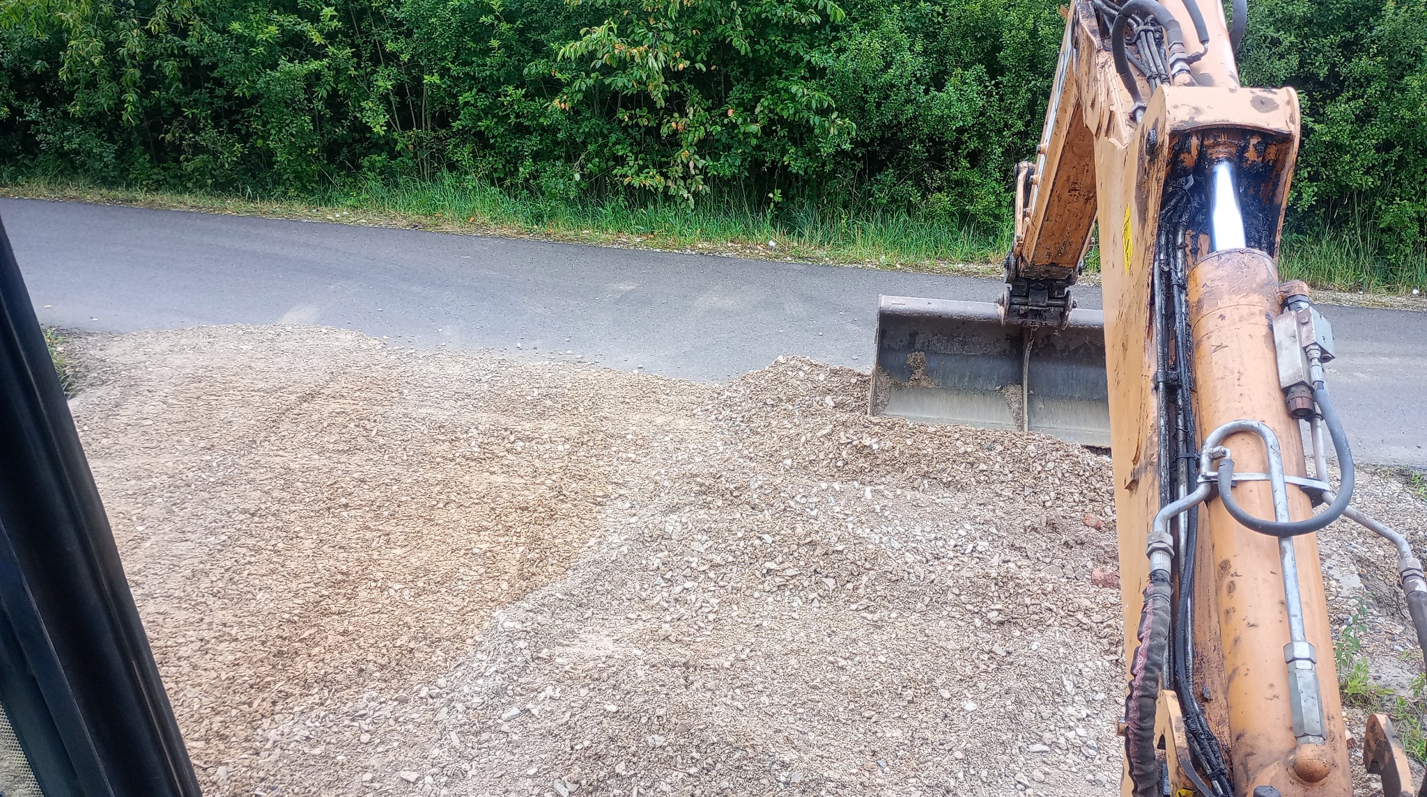 Widok z kabiny koparki na hałdę gruzu przy drodze. Ramię koparki w kadrze, zieleń w tle. Perspektywa operatora maszyny.