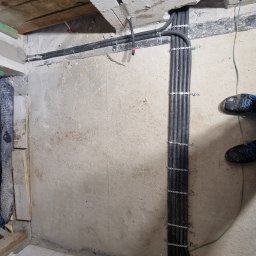 Marek Lewandowski M&M ELEKTRYKA - Wewnętrzna instalacja elektryczna: pionowy układ rur karbowanych przymocowanych do ściany, widoczne elementy sufitu i podłogi w surowym stanie.