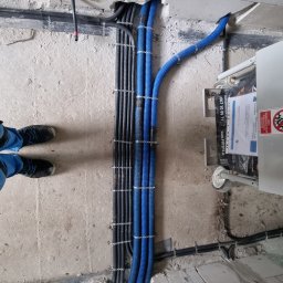 Marek Lewandowski M&M ELEKTRYKA - Rzędy czarnych i niebieskich rurek karbowanych, zamocowanych do surowej ściany. Widoczne stopy pracownika w niebieskim kombinezonie i czarnych butach roboczych.