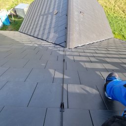 Marek Lewandowski M&M ELEKTRYKA - Szary dach z zamontowaną instalacją odgromową, widok z góry. Fragment buta w prawym dolnym rogu. W tle trawa i blaszana budka.