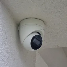 Kamera kopułkowa CCTV zamontowana na szorstkiej, jasnej ścianie. Widoczny obiektyw kamery i część obudowy. Kąt ujęcia z dołu, podkreślający dyskretny montaż.
