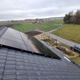 Marek Lewandowski M&M ELEKTRYKA - Panele słoneczne, czarne, na dachu z szarej dachówki, widok z góry na wiejski krajobraz w pochmurny dzień. Instalacja fotowoltaiczna na ukończonym dachu.