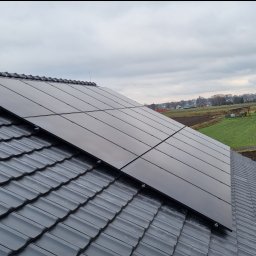 Marek Lewandowski M&M ELEKTRYKA - Panele słoneczne na dachu w Jakubowicach. Widok na cztery panele fotowoltaiczne zamontowane na szarych dachówkach. W tle pola i zabudowania.