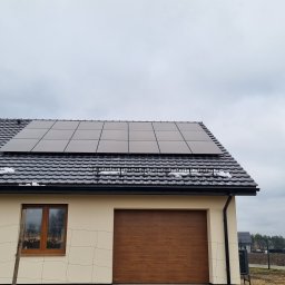 Marek Lewandowski M&M ELEKTRYKA - Panele słoneczne na dachu domu jednorodzinnego z brązową bramą garażową i oknem. Fragmenty śniegu na dachu. Pochmurne niebo w tle.