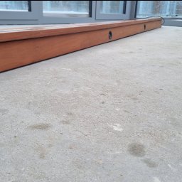 Marek Lewandowski M&M ELEKTRYKA - Drewniana listwa przypodłogowa z otworami na oświetlenie punktowe, zamontowana wzdłuż okna, na szarej, betonowej podłodze.