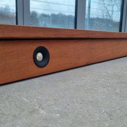 Marek Lewandowski M&M ELEKTRYKA - Drewniana listwa przypodłogowa z wbudowanymi, okrągłymi punktami świetlnymi LED o ciepłej barwie, zamontowana wzdłuż okna.