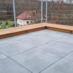 Marek Lewandowski M&M ELEKTRYKA - Nowoczesny balkon z szarymi płytkami i drewnianą balustradą. W balustradzie wbudowane są małe, okrągłe lampki oświetlające przestrzeń.