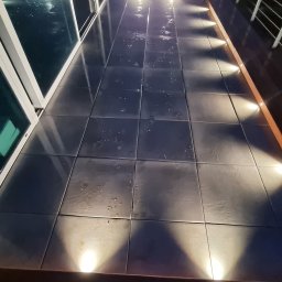 Marek Lewandowski M&M ELEKTRYKA - Podświetlony taras z płytkami i barierką, widok z góry na oświetlenie punktowe. Nocne ujęcie z butami w kadrze.