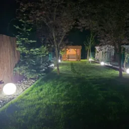 Oświetlony ogród nocą z kulistymi lampami wzdłuż ścieżki prowadzącej do altany. Światło podkreśla zieleń trawy i dekoracyjne elementy ogrodu.