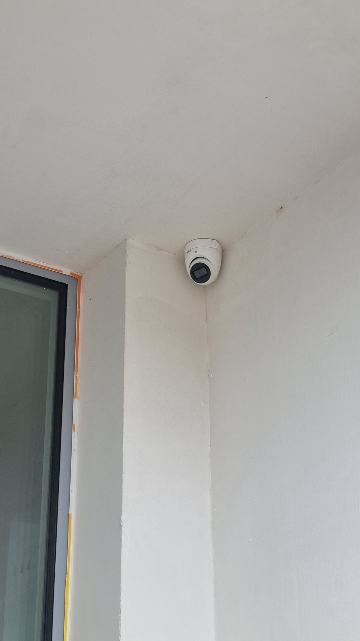 Kamera kopułkowa CCTV zamontowana w rogu pomieszczenia, widoczna obok ramy okiennej z pomarańczowym zabezpieczeniem. Białe ściany i sufit.