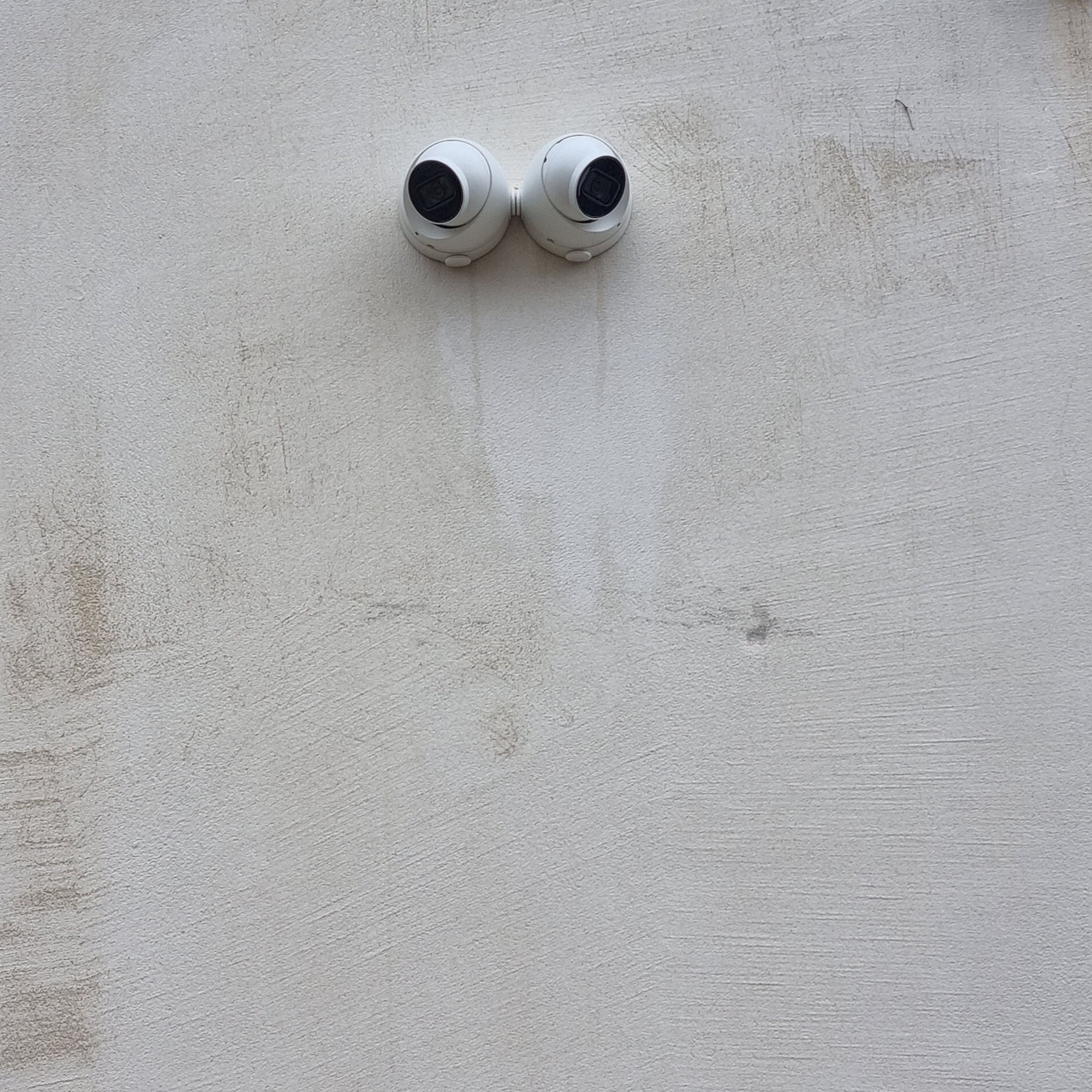 Dwie białe kamery kopułkowe CCTV zamontowane obok siebie na jasnej, teksturowanej ścianie. Kamery skierowane do przodu, z widocznymi obiektywami. Ściana z szarymi smugami.