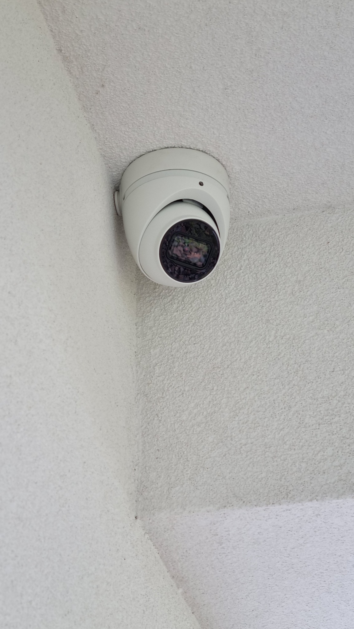 Kopułkowa kamera monitoringu, zamontowana w rogu pomieszczenia na białym, teksturowanym suficie. Widoczny obiektyw kamery. Ujęcie od dołu.