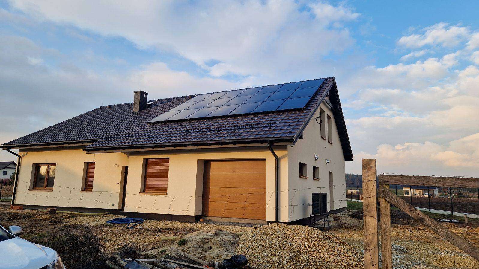 Nowoczesny dom jednorodzinny z panelami słonecznymi na dachu, brązowa dachówka, jasna elewacja, widok z zewnątrz w słoneczny dzień. Częściowo widoczny samochód.