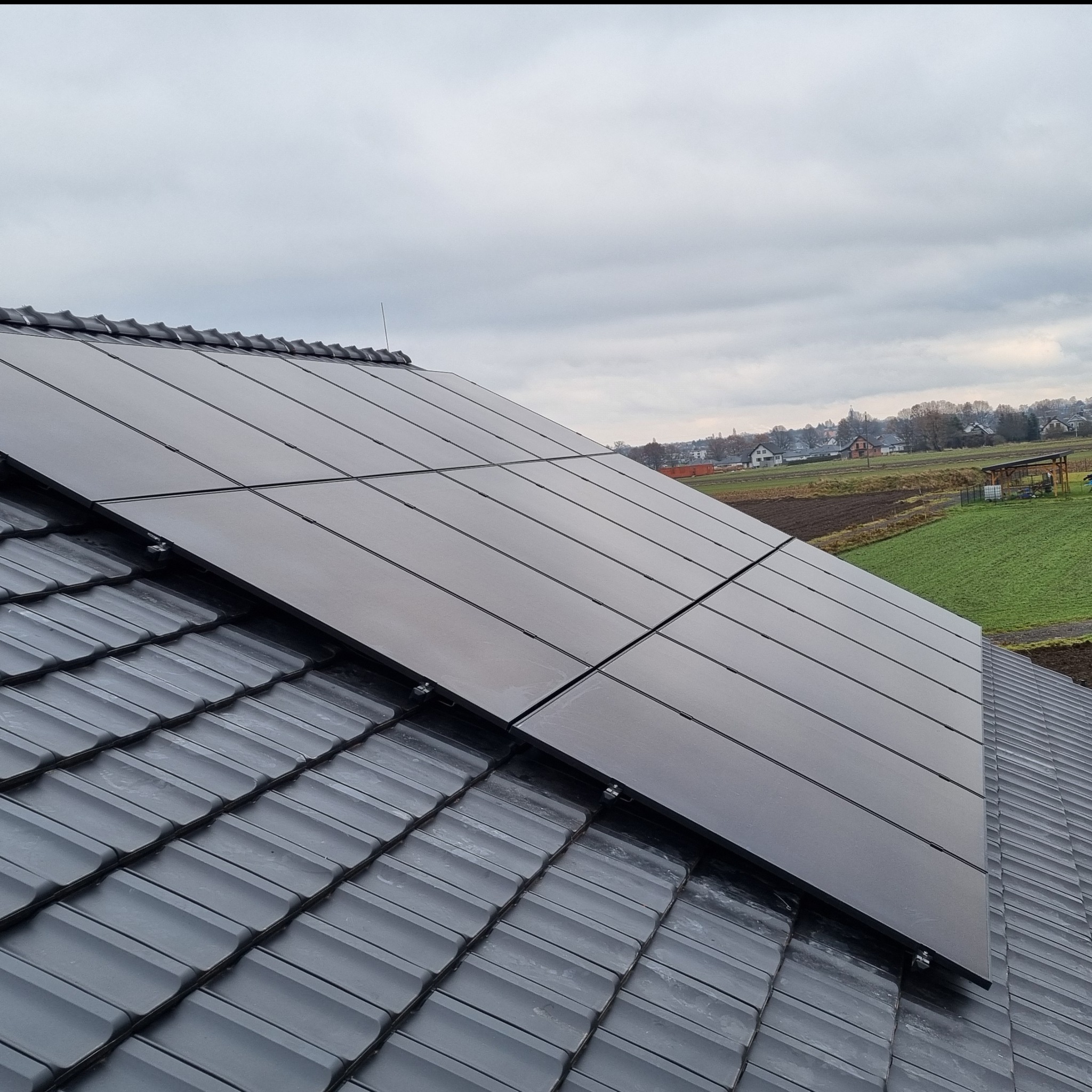 Panele słoneczne na dachu w Jakubowicach. Widok na cztery panele fotowoltaiczne zamontowane na szarych dachówkach. W tle pola i zabudowania.