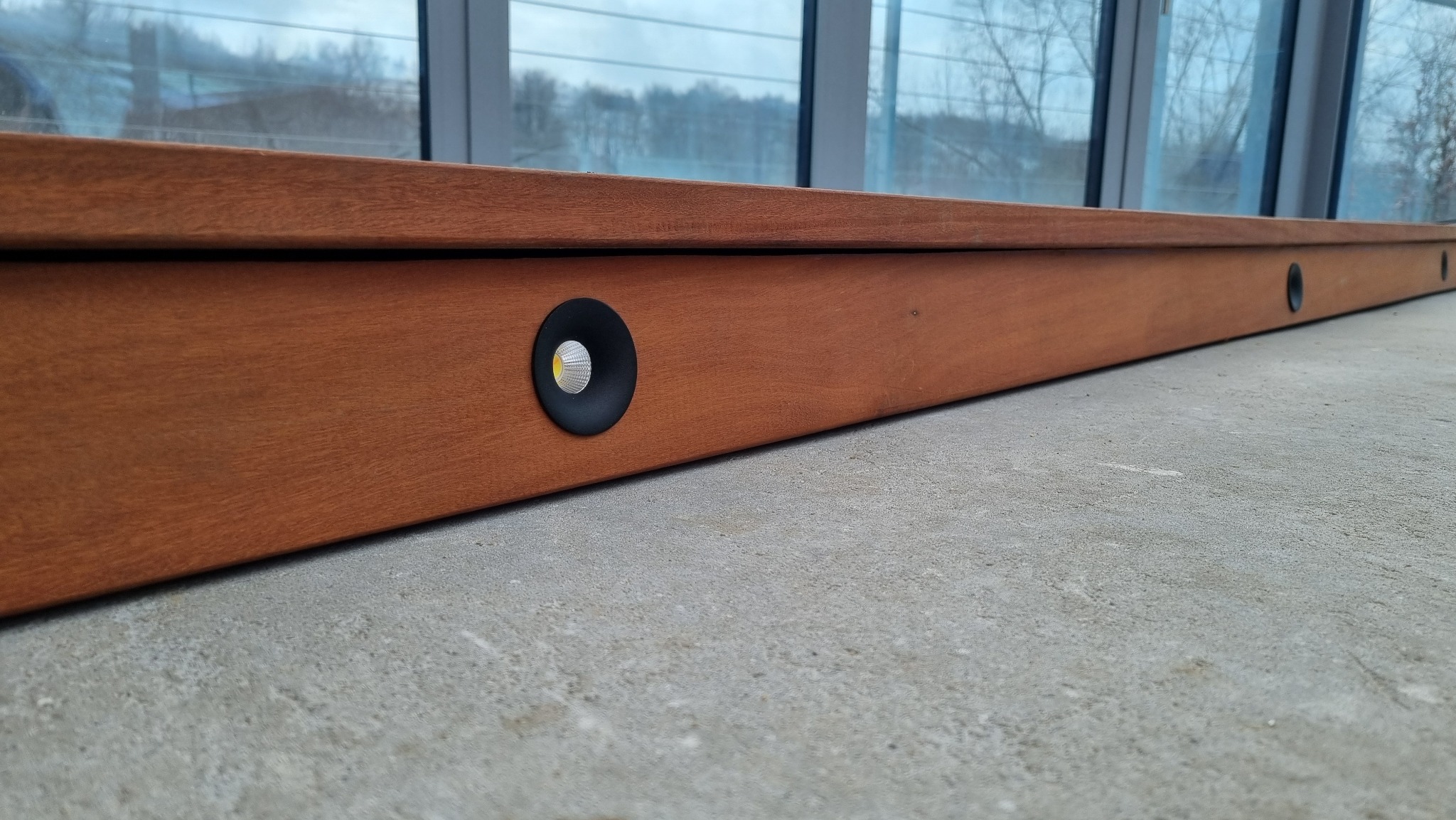 Drewniana listwa przypodłogowa z wbudowanymi, okrągłymi punktami świetlnymi LED o ciepłej barwie, zamontowana wzdłuż okna.