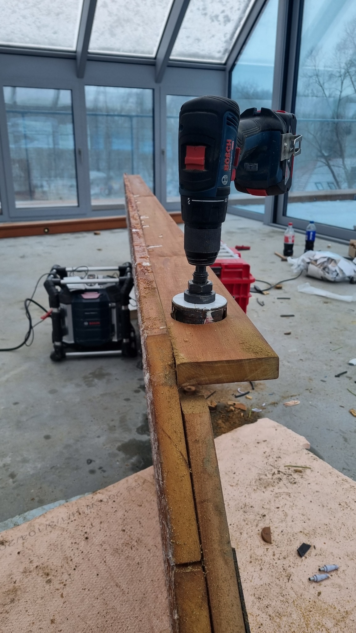 Wiertarka akumulatorowa Bosch z otwornicą wierci otwór w drewnianej belce w oranżerii. W tle widoczne narzędzia i okna.