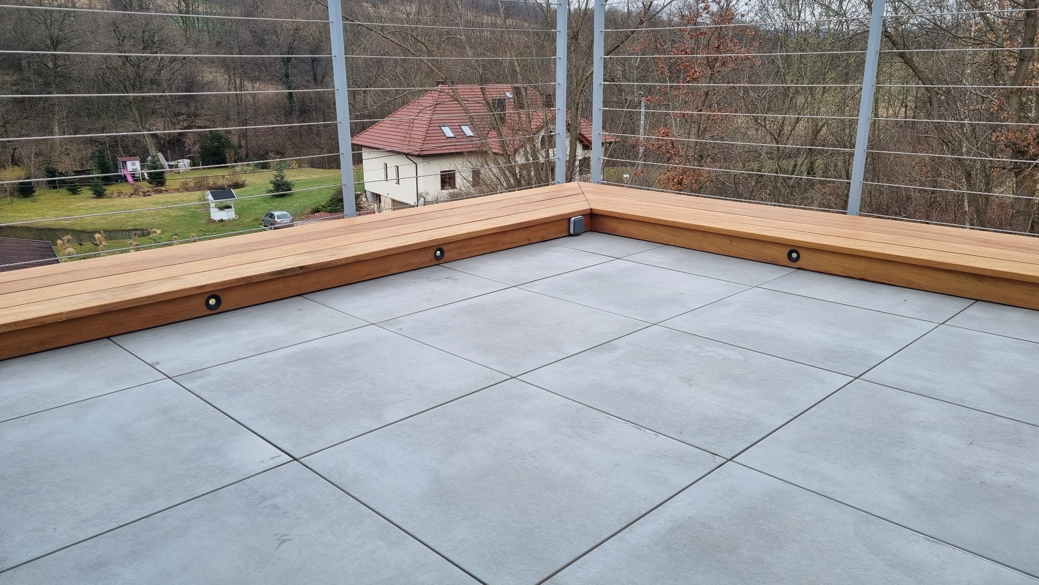 Nowoczesny balkon z szarymi płytkami i drewnianą balustradą. W balustradzie wbudowane są małe, okrągłe lampki oświetlające przestrzeń.