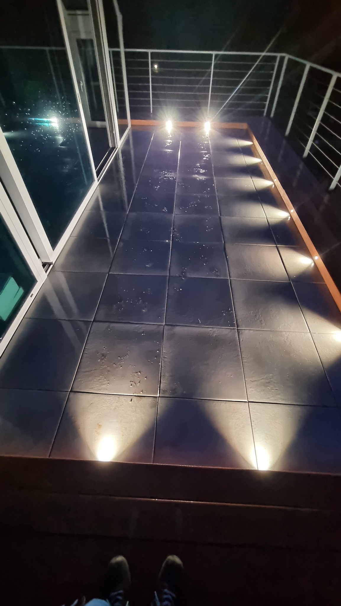 Podświetlony taras z płytkami i barierką, widok z góry na oświetlenie punktowe. Nocne ujęcie z butami w kadrze.