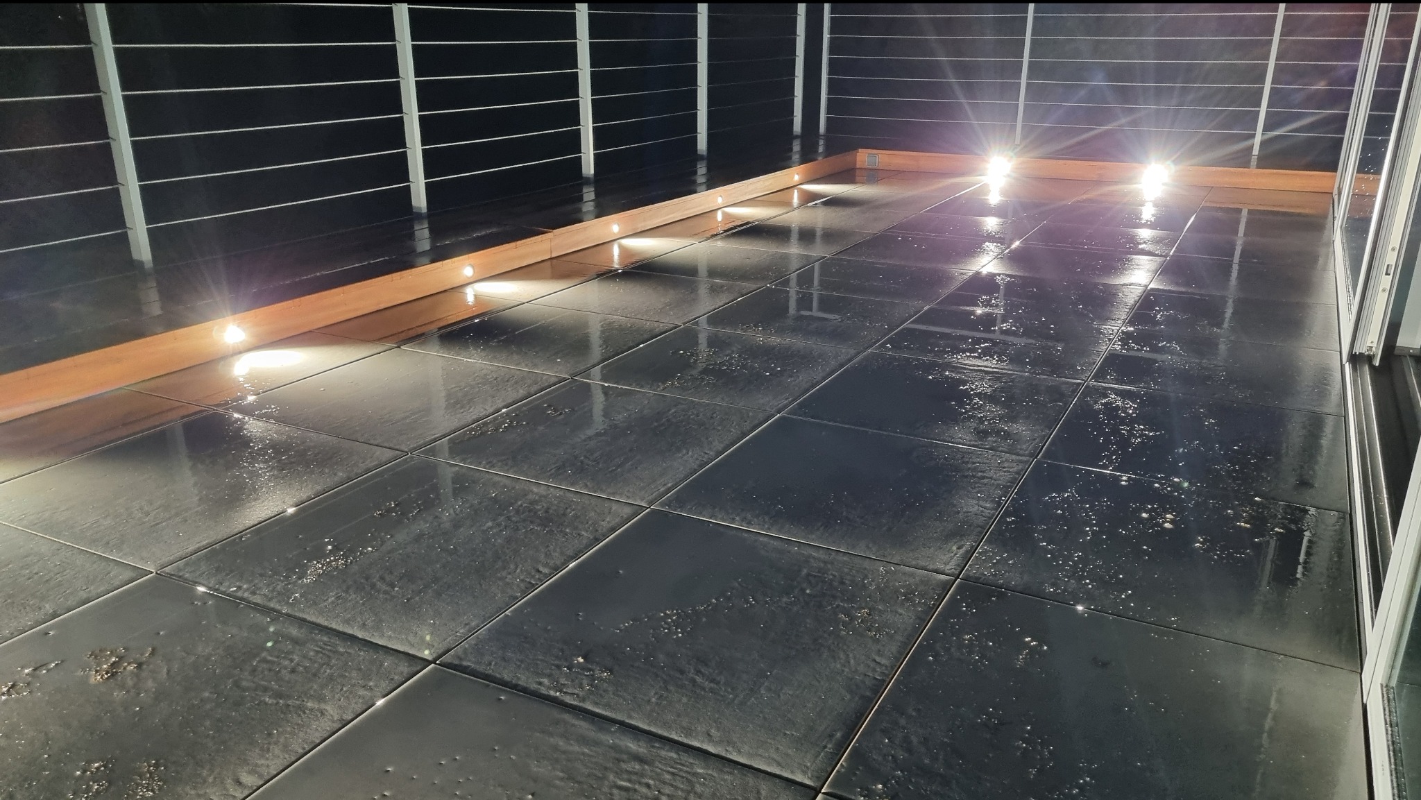 Nowoczesny balkon z czarną, błyszczącą podłogą z płytek, oświetlony punktowymi lampkami LED wbudowanymi w drewnianą listwę wzdłuż balustrady.