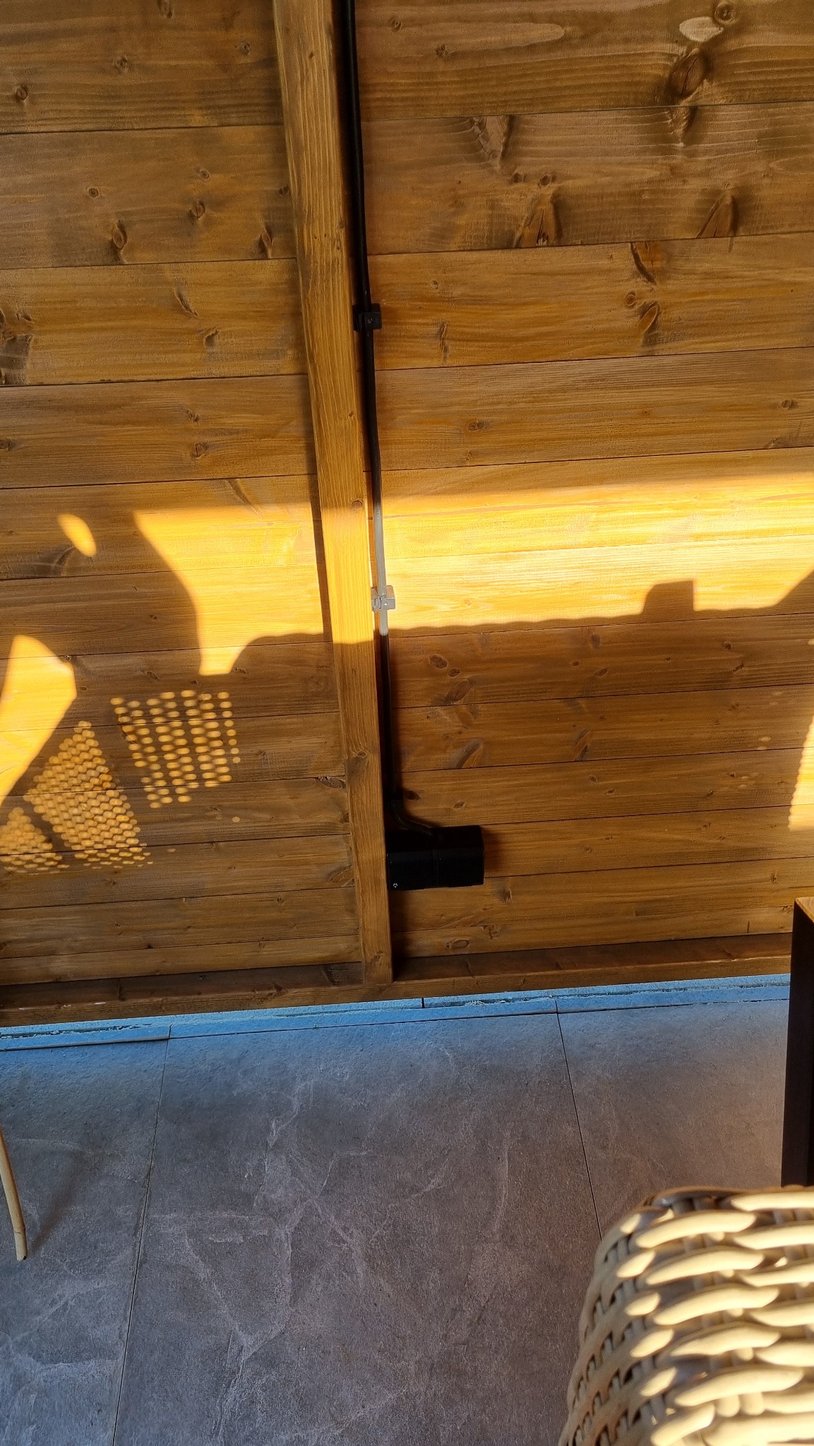 Drewniana ściana z widoczną instalacją elektryczną w czarnej rurze. Na ścianie cień ażurowego elementu. Część podłogi z szarych płytek. Ujęcie z dołu.