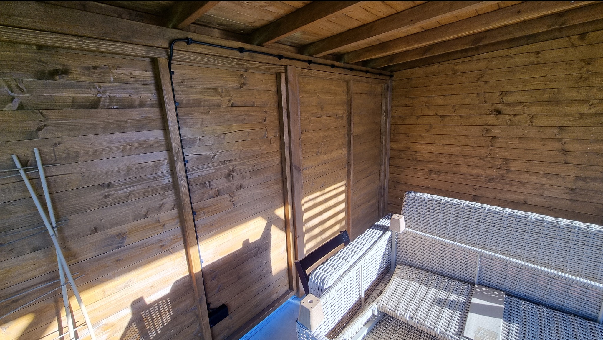 Drewniana altana z rattanowym zestawem wypoczynkowym, widoczna czarna instalacja elektryczna biegnąca po ścianie. Ujęcie z niskiej perspektywy, oświetlone promieniami słońca.