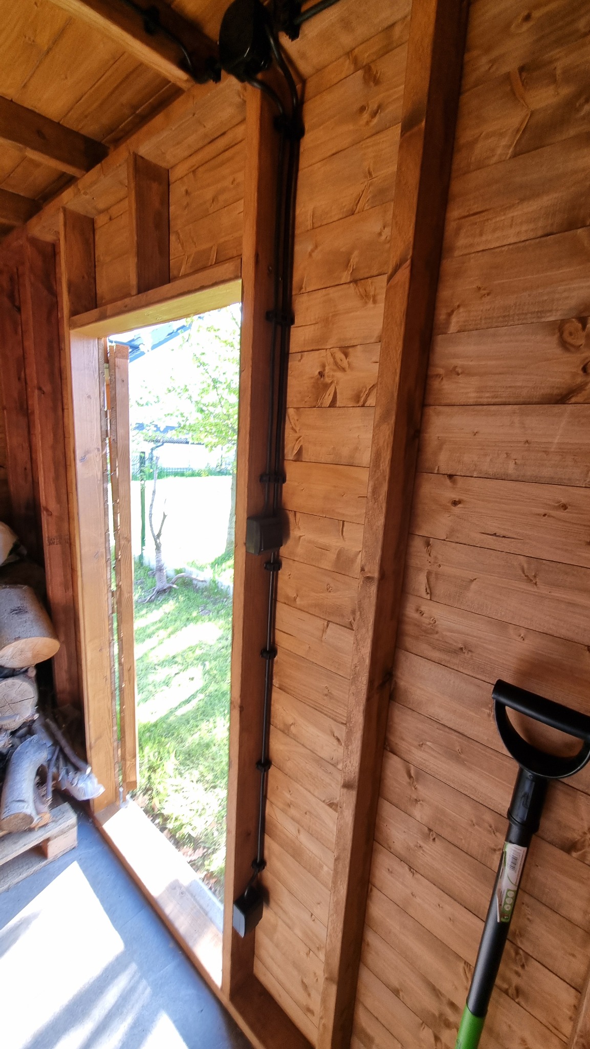 Wnętrze drewnianej konstrukcji z widoczną instalacją elektryczną poprowadzoną wzdłuż ściany. Na podłodze leżą kawałki drewna, a obok ściany oparta jest łopata.