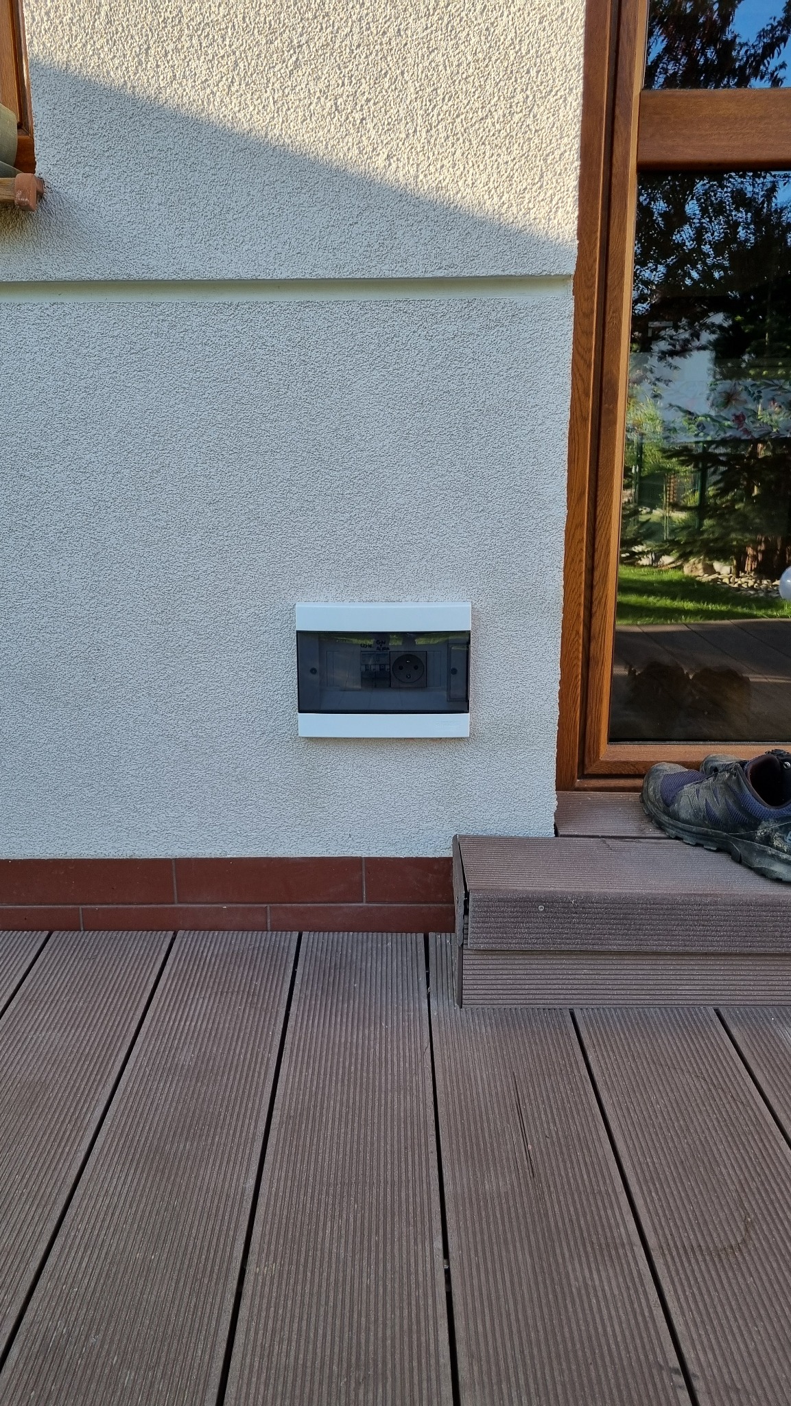 Zewnętrzna skrzynka elektryczna z gniazdem, zamontowana na elewacji domu obok okna. Widoczne buty robocze na podeście i drewniana podłoga tarasu.