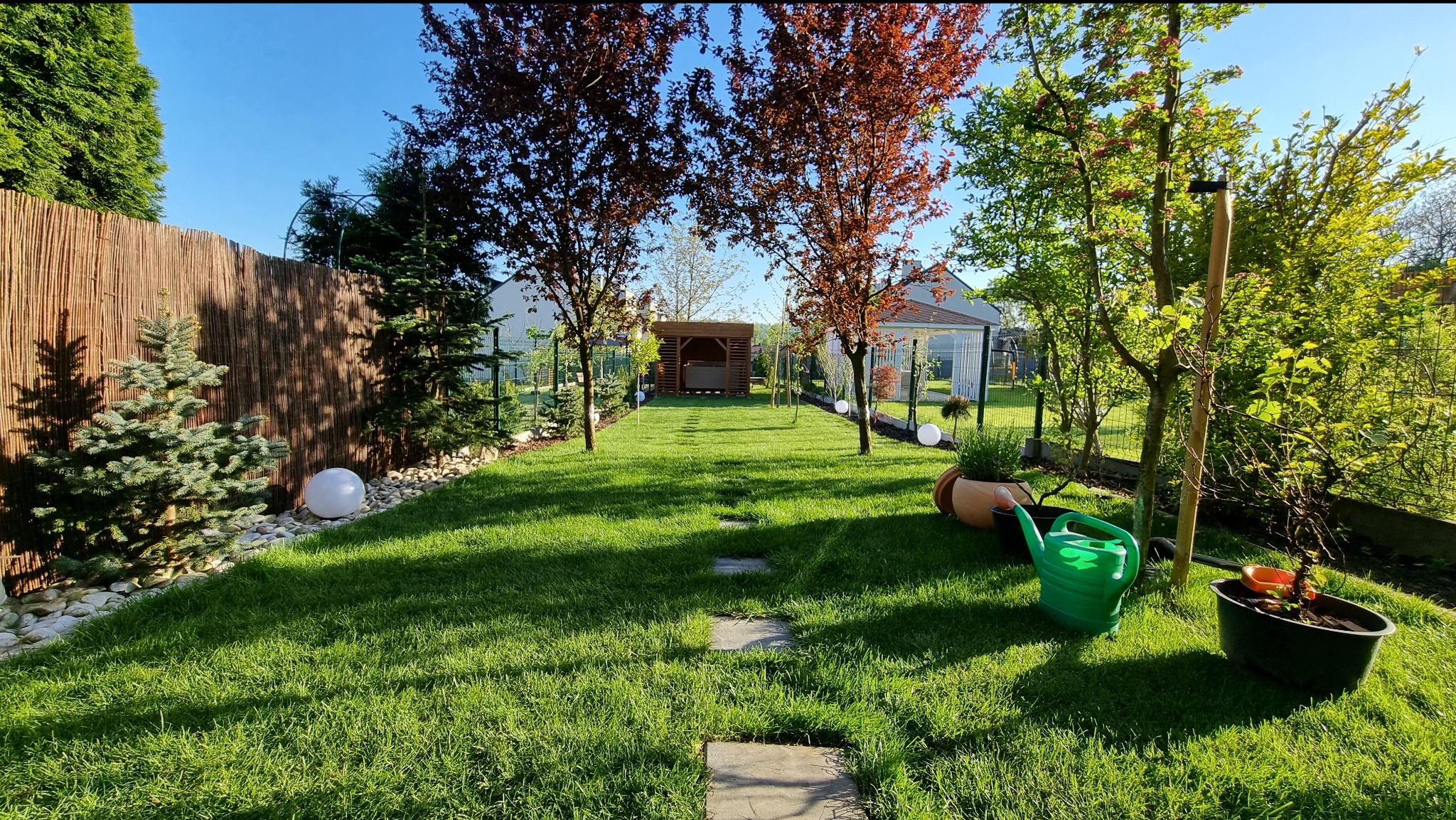 Zadbany ogród z zielonym trawnikiem, ścieżką z płyt, altaną w głębi i dekoracyjnymi kulami oświetleniowymi wzdłuż ogrodzenia. Widoczny system nawadniania.