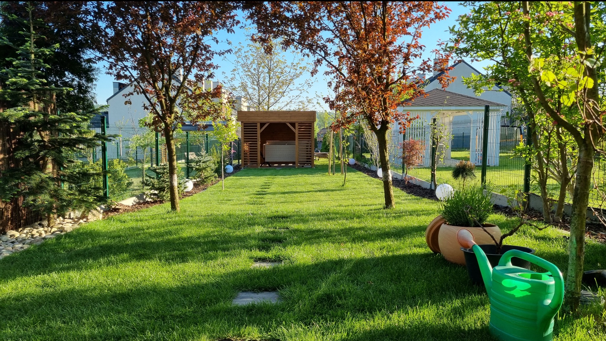 Zadbany ogród z zielonym trawnikiem, drewnianą altaną i dekoracyjnymi drzewami. Widoczny system oświetlenia, ogrodzenie i elementy małej architektury.