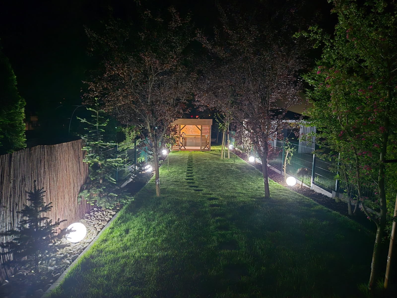 Ogród nocą z oświetleniem: ścieżka z kamieni prowadzi do altany, oświetlona kulami. Widok na zielony trawnik, drzewa i ogrodzenie. Nastrojowe, ciepłe światło.