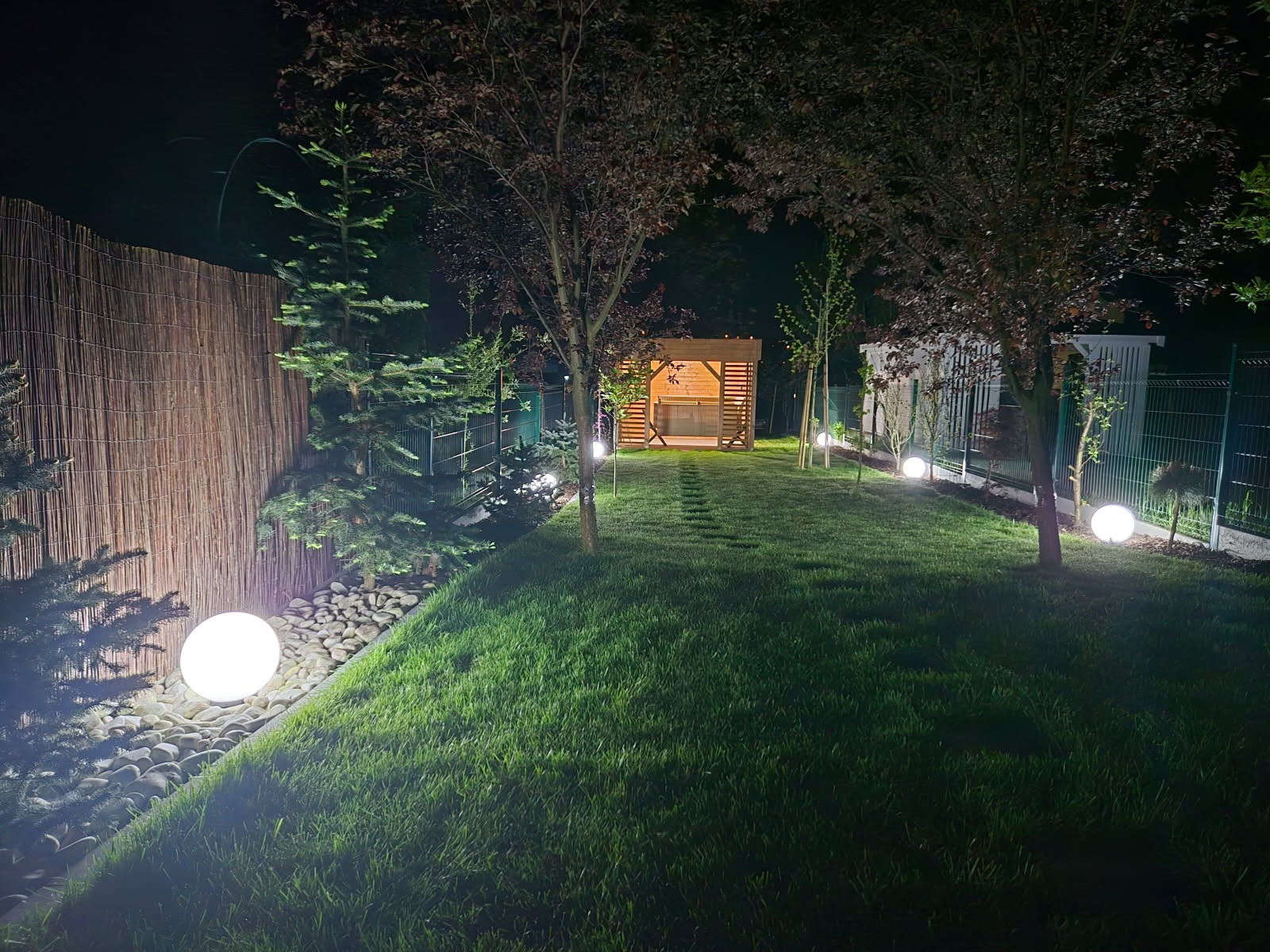 Oświetlony ogród nocą z kulistymi lampami wzdłuż ścieżki prowadzącej do altany. Światło podkreśla zieleń trawy i dekoracyjne elementy ogrodu.