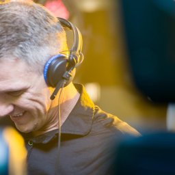 DJ Tomek Siko - Uśmiechnięty DJ w czarnej koszuli i słuchawkach z niebieskimi akcentami, oświetlony ciepłym światłem, w trakcie występu. Widoczne elementy sprzętu.