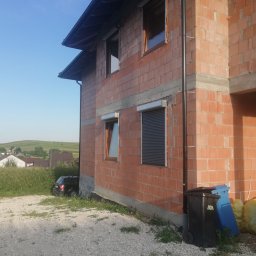 KJ SERWIS - Surowy budynek z czerwonej cegły z zamontowanymi oknami i roletami zewnętrznymi. Widoczne śmietniki i samochód w tle.