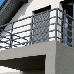 KJ SERWIS - Nowoczesny balkon z szarą, metalową balustradą na tle białej elewacji domu z oknami PCV i roletami. Minimalistyczny design i stonowana kolorystyka.