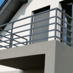 Nowoczesny balkon z szarą, metalową balustradą na tle białej elewacji domu z oknami PCV i roletami. Minimalistyczny design i stonowana kolorystyka.