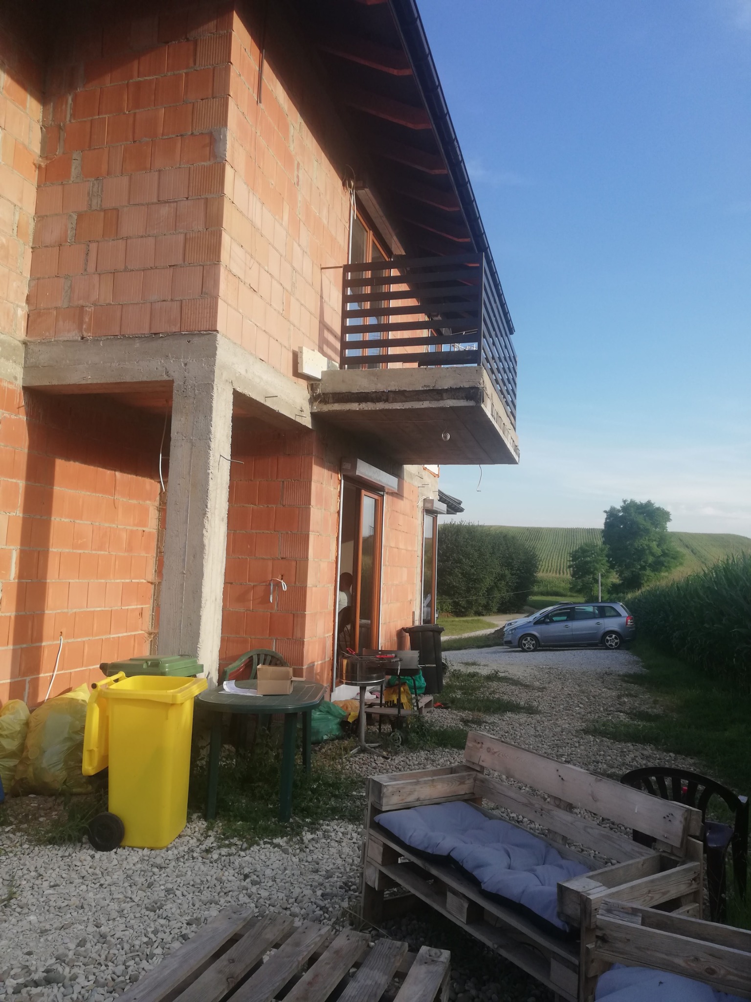Niedokończony dwukondygnacyjny dom z cegły, widoczny balkon z drewnianą balustradą, w tle pole uprawne i samochód. Na pierwszym planie palety i śmietnik.