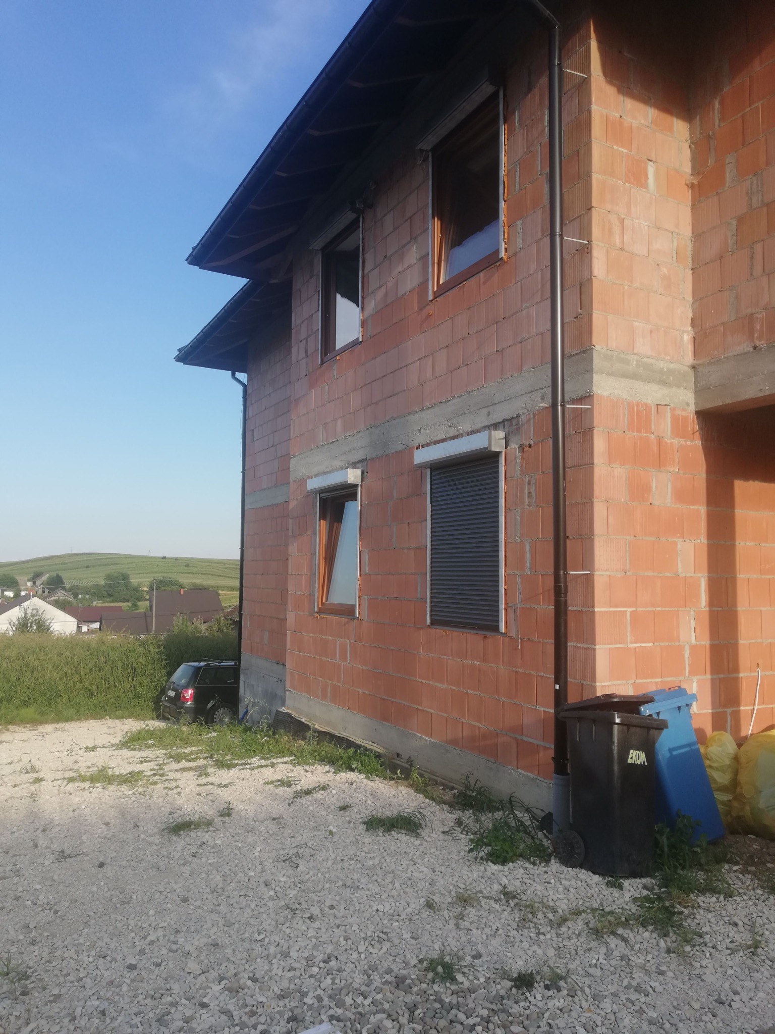 Surowy budynek z czerwonej cegły z zamontowanymi oknami i roletami zewnętrznymi. Widoczne śmietniki i samochód w tle.