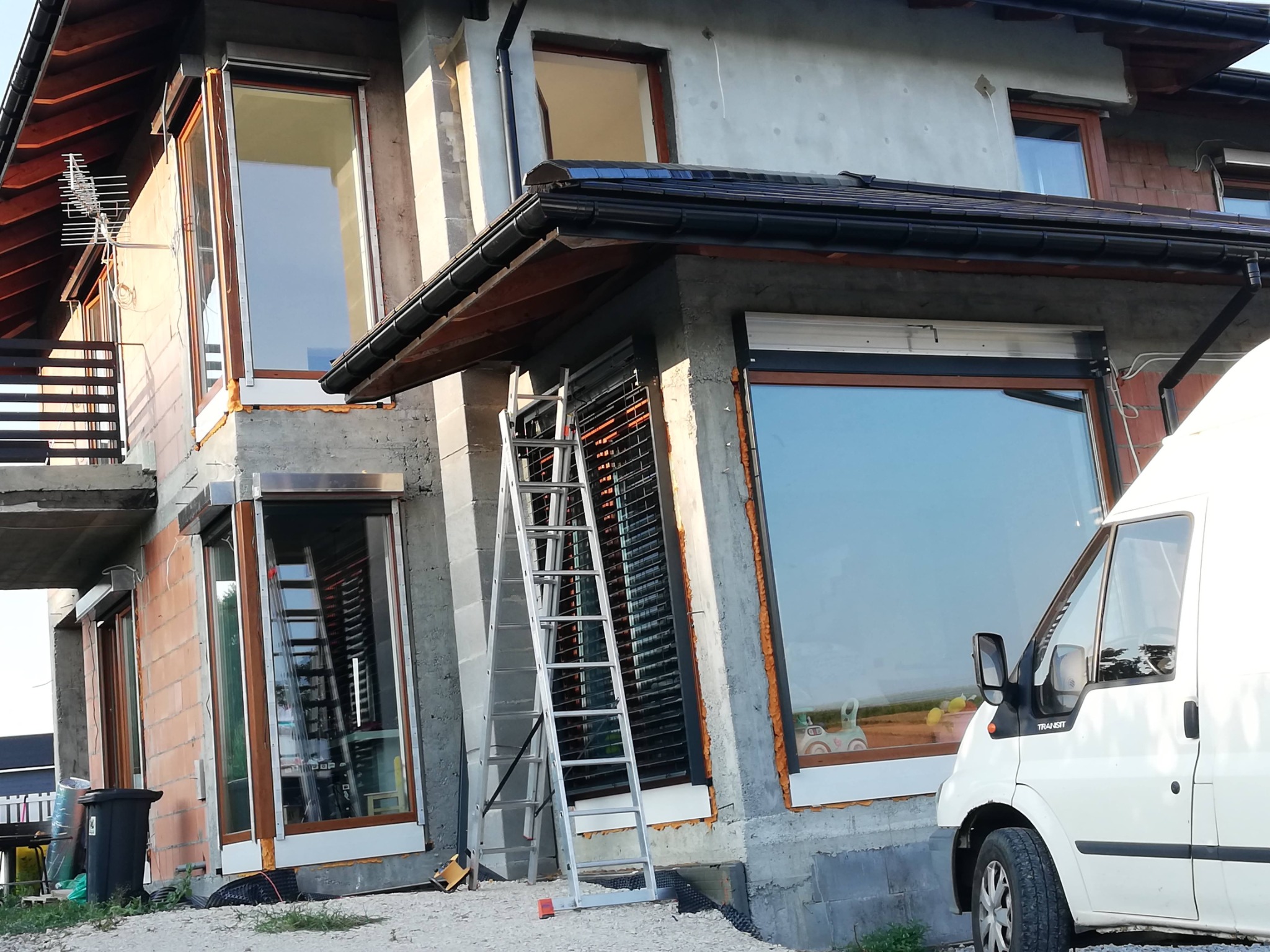 Montaż rolet zewnętrznych na oknach budynku w trakcie budowy. Widoczna drabina oparta o okno, samochód dostawczy i surowa elewacja.