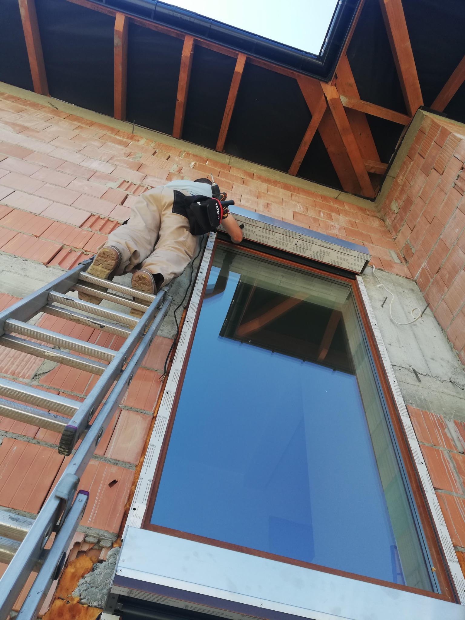 Montaż rolety zewnętrznej na oknie w surowym budynku z cegły. Pracownik na drabinie z wiertarką. Widoczna konstrukcja dachu. Ujęcie z dołu.