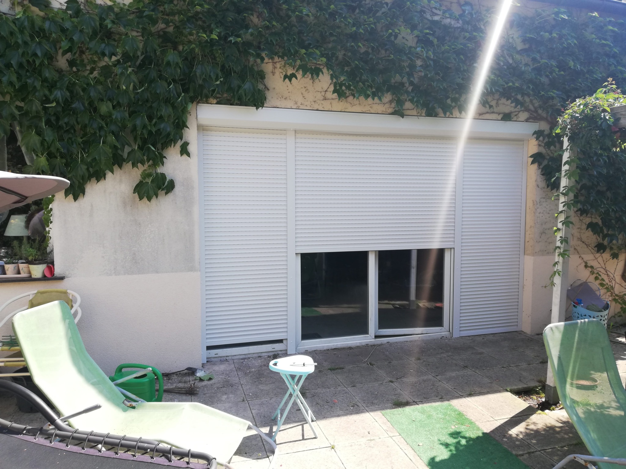 Biała roleta zewnętrzna, częściowo opuszczona, na tle ściany porośniętej bluszczem. Widoczne krzesła ogrodowe i fragment kostki brukowej.