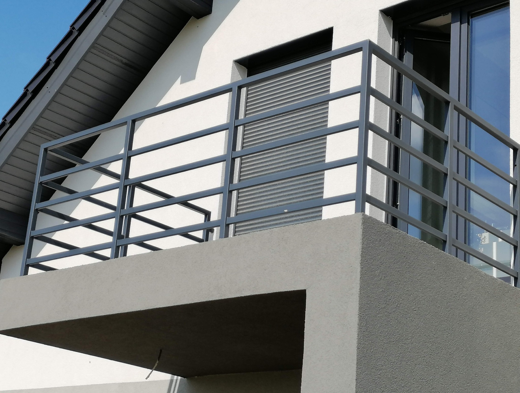 Nowoczesny balkon z szarą, metalową balustradą na tle białej elewacji domu z oknami PCV i roletami. Minimalistyczny design i stonowana kolorystyka.