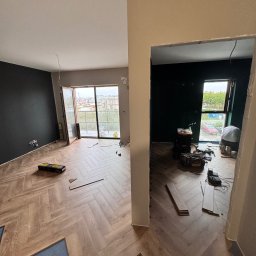 PreciseHomeRenowation - Układanie parkietu w jodełkę francuską w mieszkaniu w Poznaniu. Widoczne narzędzia i częściowo ułożona podłoga. Ściany w kolorach szarości i zieleni.