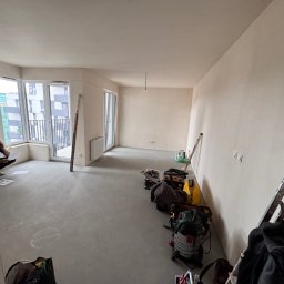 PreciseHomeRenowation - Wnętrze w trakcie prac wykończeniowych: gładkie ściany, surowa podłoga, narzędzia i drabina. Mężczyzna analizuje dokumenty przy oknie balkonowym.