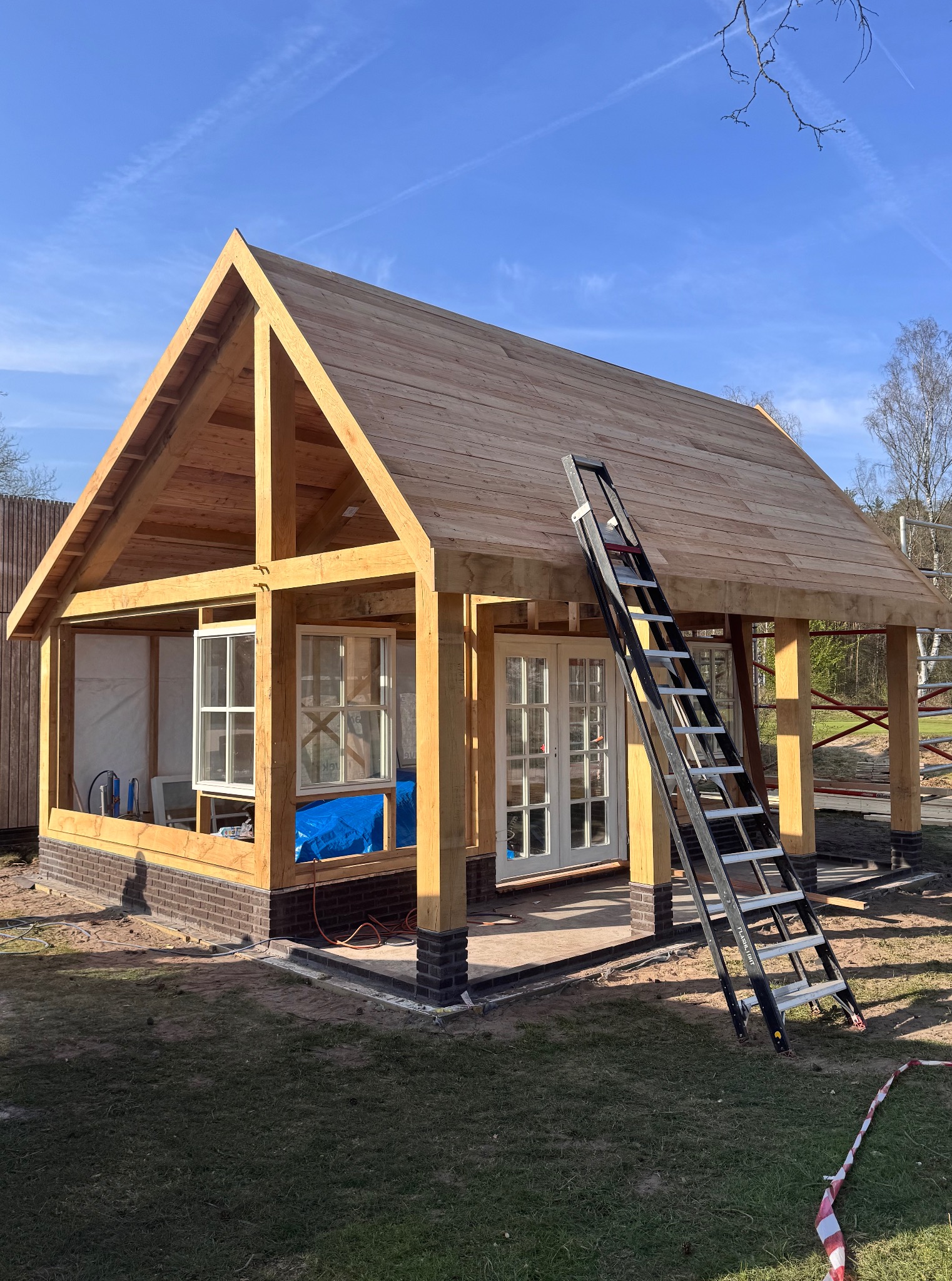 Drewniana konstrukcja altany w trakcie budowy, widoczne elementy więźby dachowej, okna, drzwi i drabina oparta o budowlę na tle błękitnego nieba.