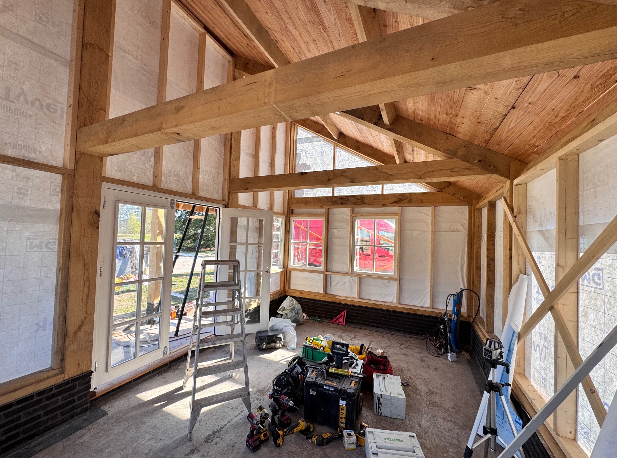 Wnętrze w trakcie budowy z drewnianą konstrukcją szkieletową, oknami i drzwiami. Widoczne narzędzia i drabina sugerują trwające prace wykończeniowe.